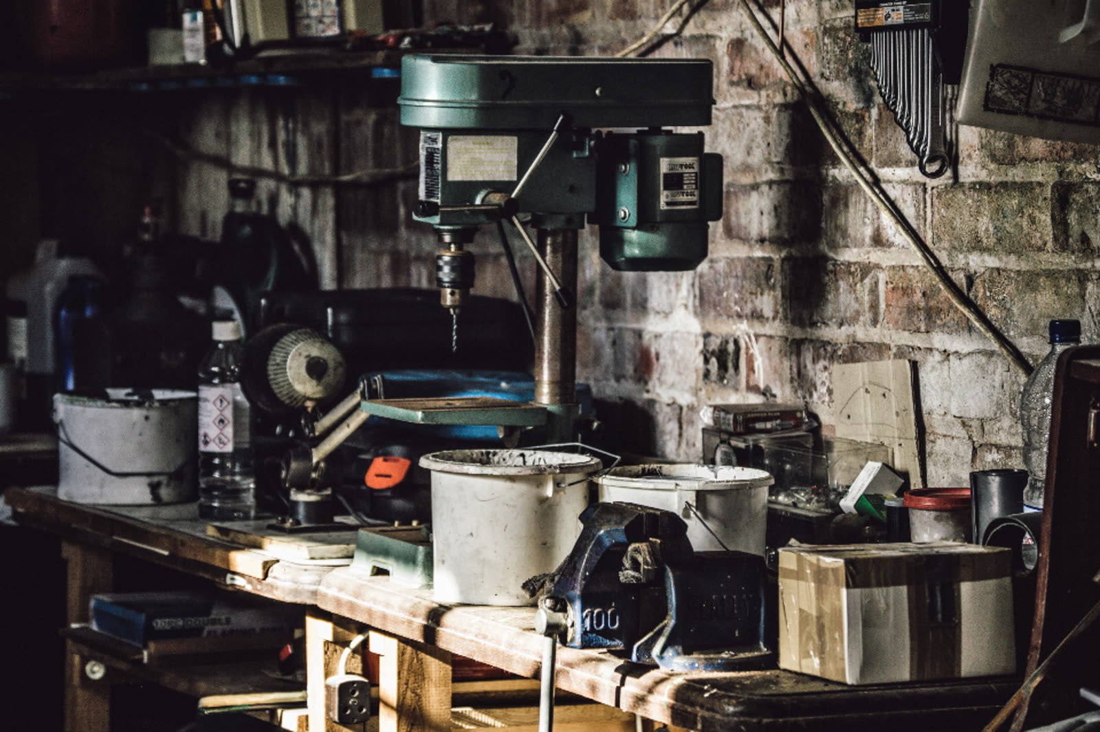 A workshop bench against a brick wall, with a drill press, clamps and hand tools laid out within reach.